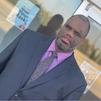 A person wearing a dark blue suit jacket, a light purple collared shirt, and a patterned tie standing outside a building with large glass windows. The person has short dark hair, a graying beard, and is looking slightly to the left with their eyes partially closed. A sign on the window in the upper left corner reads "Wear Your Mask!" in blue lettering.